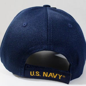 May include: Navy blue baseball cap with a curved brim. The adjustable strap features the text "U.S. NAVY" in gold lettering. The cap is made of a durable fabric and has a button on top.