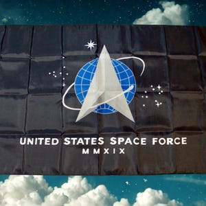 Op de afbeelding: Zwarte vlag met het embleem van de United States Space Force. Het embleem bevat een witte ster, een gestileerde witte pijl en een blauwe wereldbol met witte lijnen. De tekst "UNITED STATES SPACE FORCE MMXIX" is wit gedrukt.