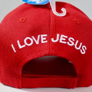 May include: Red baseball cap with the embroidered text "I LOVE JESUS" in white. The cap has a curved brim and a classic six-panel design. A white plastic tag holder is attached to the cap.