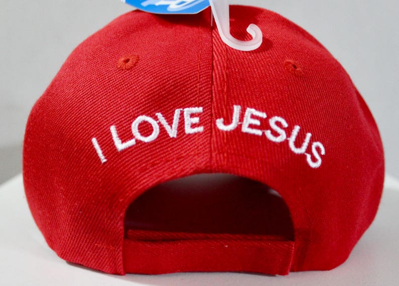 May include: Red baseball cap with the embroidered text "I LOVE JESUS" in white. The cap has a curved brim and a classic six-panel design. A white plastic tag holder is attached to the cap.