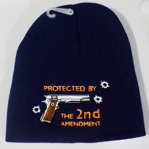 May include: Navy blue beanie with the embroidered text "PROTECTED BY THE 2ND AMENDMENT" and a pistol graphic. The design includes bullet hole details. The beanie has a plastic hang tag attached.
