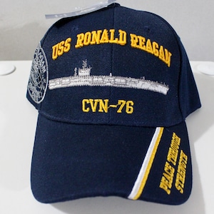 May include: Navy blue baseball cap with yellow embroidered text "USS RONALD REAGAN" and "CVN-76". The cap features a white and yellow stripe on the brim and the words "PEACE THROUGH STRENGTH". A patch is on the side.