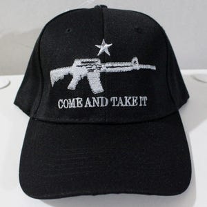 Op de afbeelding: Zwarte baseballcap met een wit geborduurd geweer, ster en de tekst "COME AND TAKE IT". De cap heeft een gebogen klep en een gestructureerde kroon. Een klassiek, casual accessoire.