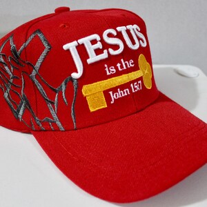 May include: A red baseball cap with the text "JESUS is the John 15:7" embroidered in white and yellow. The cap features a stylized cross design on the side. The cap has a curved brim and a button on top.