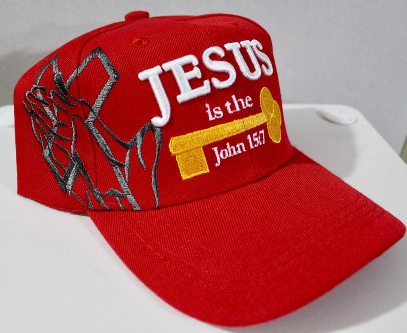 May include: A red baseball cap with the text "JESUS is the John 15:7" embroidered in white and yellow. The cap features a stylized cross design on the side. The cap has a curved brim and a button on top.