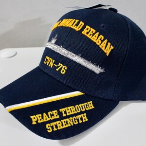 May include: Navy blue baseball cap with embroidered text and a ship design. The cap features the words "Ronald Reagan", "CVN-76", and "Peace Through Strength" in yellow. A white and yellow stripe accents the brim.