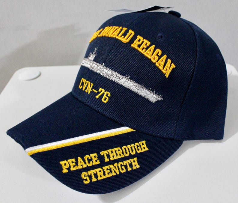 May include: Navy blue baseball cap with embroidered text and a ship design. The cap features the words "Ronald Reagan", "CVN-76", and "Peace Through Strength" in yellow. A white and yellow stripe accents the brim.