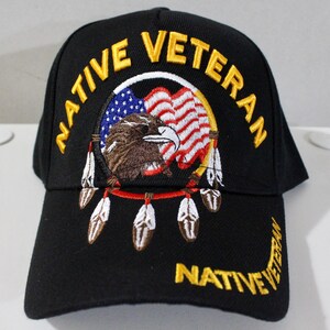 Può includere: Un cappellino da baseball nero con il testo giallo ricamato NATIVE VETERAN. Il centro presenta un design circolare acchiappasogni con una testa d'aquila marrone, un motivo a bandiera americana rossa, bianca e blu e piume bianche.