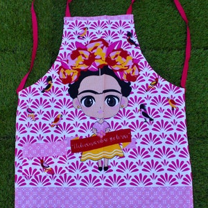 May include: Pink and white kitchen apron featuring a cartoon image of Frida Kahlo wearing a flower crown and holding a paintbrush. The apron has a pink pocket with the text "Pinté mis sueños y vivieron" in Spanish.