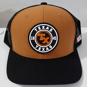 May include: A brown and black trucker cap with a curved peak. The cap features a circular patch with the text "TEXAS" and a "Tx" logo. The cap has a black mesh back and a snap closure.