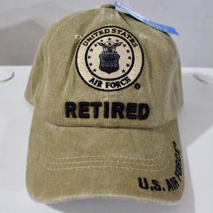 May include: A tan baseball cap with the words "RETIRED" embroidered in black across the front. The cap features a circular patch with the words "UNITED STATES AIR FORCE" and an emblem. The text "U.S. AIR FORCE" is embroidered on the side.