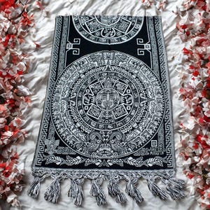 May include: Black and white Aztec calendar design scarf with fringe. The scarf features a detailed depiction of the Aztec calendar, with intricate patterns and symbols. The scarf is rectangular and has a fringe along the bottom edge.