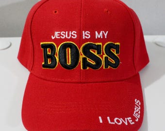 May include: A red baseball cap with the text "JESUS IS MY BOSS" embroidered in white and black. The phrase "I LOVE JESUS" is also embroidered on the brim. The cap has a curved brim and a button on top.