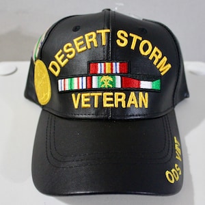 Puede incluir: Gorra de béisbol negra con bordado amarillo "DESERT STORM VETERAN". Presenta cintas militares y un parche de medalla dorada. La visera tiene bordado amarillo "Ods Vet".