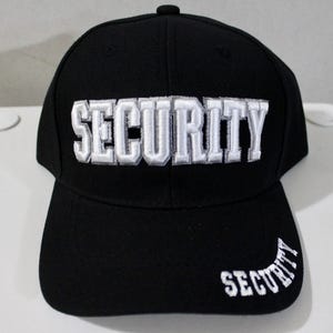May include: A black baseball cap featuring the word SECURITY embroidered in bold white capital letters across the front panel. A smaller version of the word SECURITY is also embroidered in white on the right side of the curved brim.