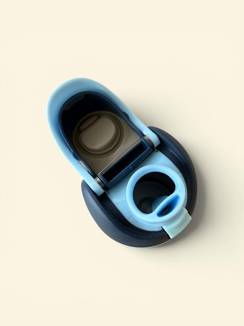 May include: A close-up of a blue and navy water bottle lid. The lid is open, revealing the interior. The lid has a light blue spout and a dark blue base. The lid is made of plastic.