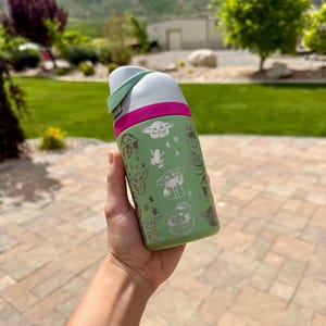 May include: A light green water bottle featuring a repeating white graphic pattern of a small creature with large ears, frogs, and stars. The bottle has a bright pink band around the top and a white lid with a light green accent piece on the side.