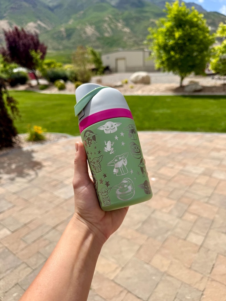 May include: A light green water bottle featuring a repeating white graphic pattern of a small creature with large ears, frogs, and stars. The bottle has a bright pink band around the top and a white lid with a light green accent piece on the side.