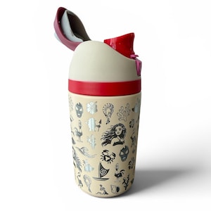 May include: A cream-colored travel mug with a red lid and accents. The mug is decorated with various black and silver illustrations, including flowers, faces, and sea creatures. The lid is open, revealing a white interior.