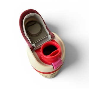 May include: An open, cream-colored travel mug with a red interior and accents. The lid is open, revealing the drinking area and a stainless steel hinge. The mug has a small pink tab and a decorative design at the base.
