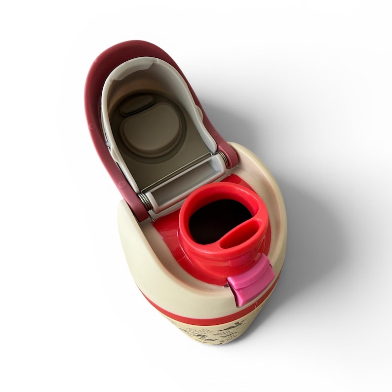 May include: An open, cream-colored travel mug with a red interior and accents. The lid is open, revealing the drinking area and a stainless steel hinge. The mug has a small pink tab and a decorative design at the base.