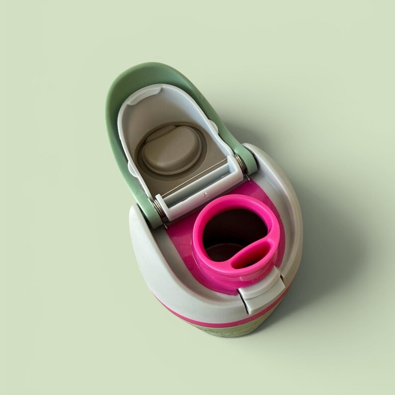 May include: A water bottle lid featuring a sage green flip-top cap with a hinge mechanism, a light grey outer shell, and a vibrant hot pink circular drinking spout insert. The internal components include a beige silicone seal and a white plastic frame.