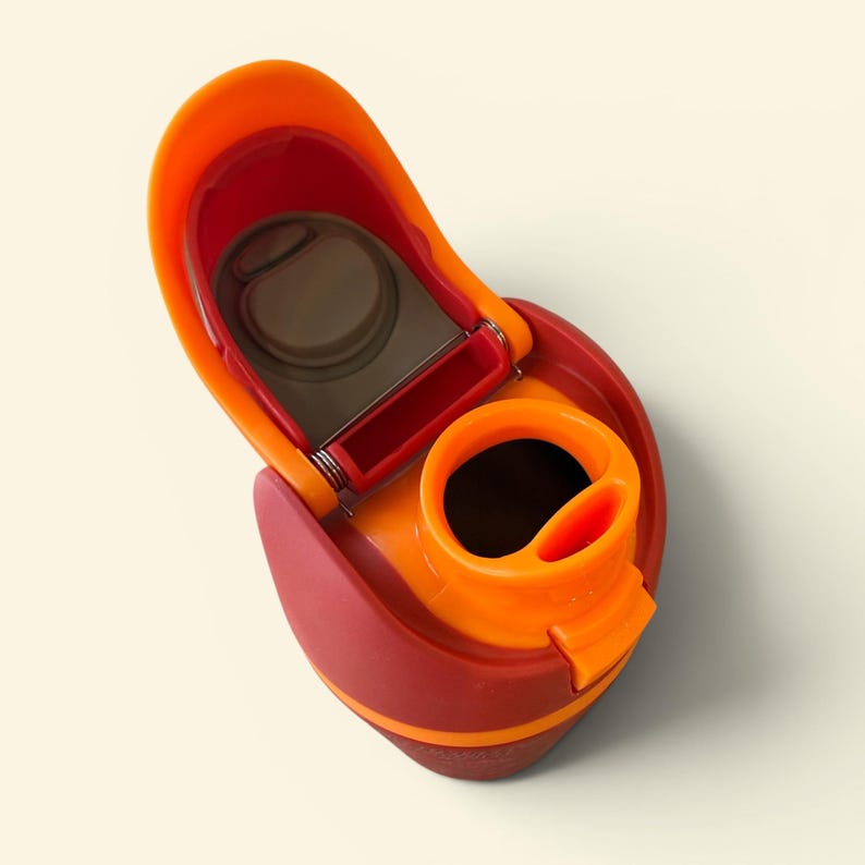 May include: A close-up of a water bottle lid in shades of orange and red. The lid is open, revealing the interior components. The design includes a flip-top opening and a drinking spout.