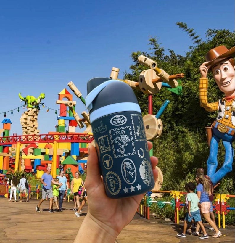 May include: A navy blue water bottle with a light blue lid and accents. The bottle features various cartoon character stamps and the word "Woody". The background shows a theme park with a Woody statue and colorful structures.