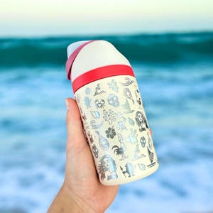 May include: Cream-colored insulated water bottle with a red band and white top. The bottle is covered in silver illustrations of sea creatures and characters. The bottle is held in front of a blurred ocean background.