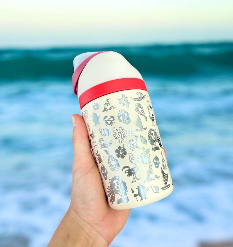 May include: Cream-colored insulated water bottle with a red band and white top. The bottle is covered in silver illustrations of sea creatures and characters. The bottle is held in front of a blurred ocean background.
