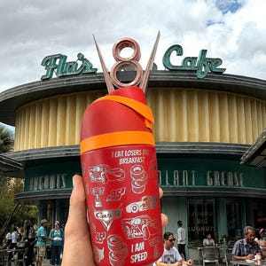 May include: A red travel mug with an orange band and a decorative top. The mug features silver illustrations of cars, tires, and the text "I EAT LOSERS FOR BREAKFAST" and "I AM SPEED". The background shows a cafe.