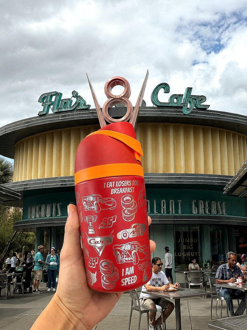 May include: A red travel mug with an orange band and a decorative top. The mug features silver illustrations of cars, tires, and the text "I EAT LOSERS FOR BREAKFAST" and "I AM SPEED". The background shows a cafe.