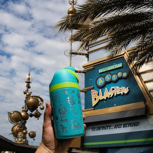May include: A teal water bottle with a green lid and a silver design featuring Buzz Lightyear and space-themed illustrations. The bottle is held in front of a sign that reads "Buzz Lightyear Astro Blasters" and "To Infinity... And Beyond!"