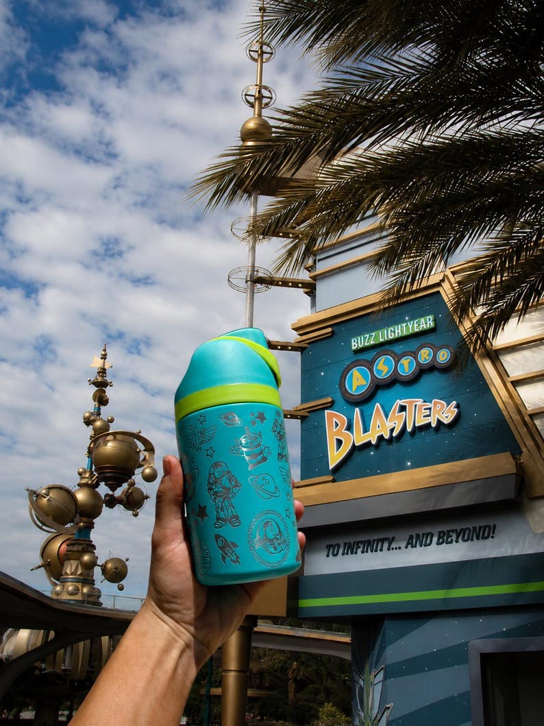 May include: A teal water bottle with a green lid and a silver design featuring Buzz Lightyear and space-themed illustrations. The bottle is held in front of a sign that reads "Buzz Lightyear Astro Blasters" and "To Infinity... And Beyond!"