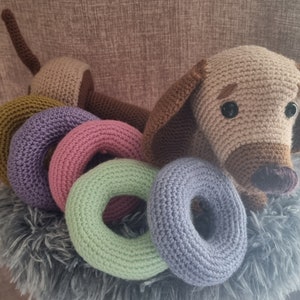 May include: A crocheted dachshund toy with brown and beige fur, sitting on a grey fluffy surface. The dachshund is surrounded by five colourful crocheted rings in shades of green, purple, pink, and brown.