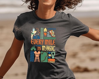 Może przedstawiać: Ciemnoszara koszulka z napisem "EVERY MILE IS MAGIC" w kolorze pomarańczowym i białym. Koszulka posiada kolorową grafikę wróżki, buta do biegania, zamku i innych ikon. Koszulka ma okrągły dekolt.