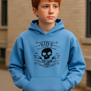 May include: A pale blue hoodie with a black skull graphic and the text "SID'S TOY CHOP SHOP est. 1995". The hoodie has a front pocket and a hood. The image shows a person wearing the hoodie.