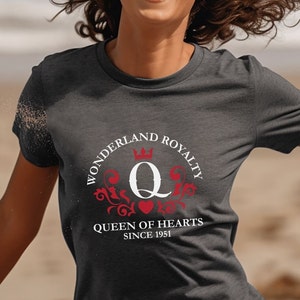 以下が含まれることがあります： 「Wonderland Royalty」と「Queen of Hearts Since 1951」の文字が入った円形デザインが特徴の濃いグレーのTシャツ。デザインには王冠、文字「Q」、赤い装飾要素が含まれています。Tシャツは半袖です。