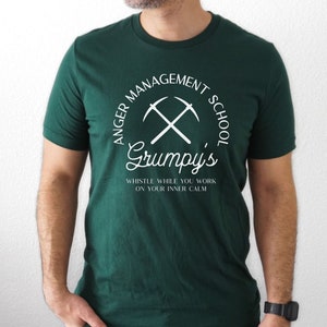 以下が含まれることがあります： 白い文字が入った濃い緑色のTシャツ。「ANGER MANAGEMENT SCHOOL」の文字が半円状に、「Grumpy's」の文字と交差したツルハシの上に配置されています。下には「Whistle while you work on your inner calm」と書かれています。