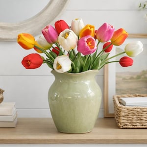 May include: A vase of vibrant tulips in a pale green ceramic vase. The tulips are in shades of red, pink, yellow, and white. The vase sits on a wooden surface, with a mirror and other decorative items visible in the background.