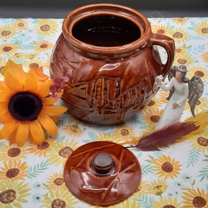 May include: A brown ceramic jar with a lid and a handle. The jar has a textured surface and is decorated with a pattern of lines and swirls. The lid is also brown and has a knob on top. The jar is sitting on a white and green floral patterned surface.