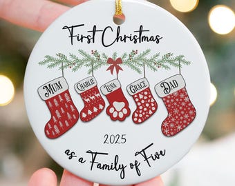 May include: A white ceramic Christmas ornament with a gold string. The ornament features the text "First Christmas" and "as a Family of Five" with the year 2025. It includes five Christmas stockings with names: Mum, Charlie, Luna, Grace, and Dad. The stockings are red and white.