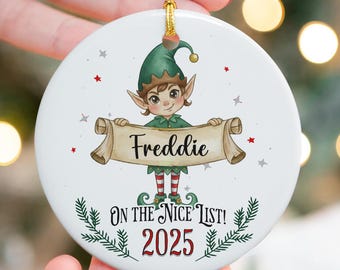 May include: A white ceramic Christmas ornament featuring a cartoon elf holding a banner with the name "Freddie." The ornament includes the text "ON THE NICE LIST! 2025" and is decorated with stars and green accents. A gold string is attached for hanging.