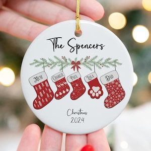 May include: White ceramic Christmas ornament with a red and white stocking design. The ornament features the text "The Spencers" and "Christmas 2024". The stockings are labeled "Mum", "Charlie", "Paul", "Lola", and "Dad".