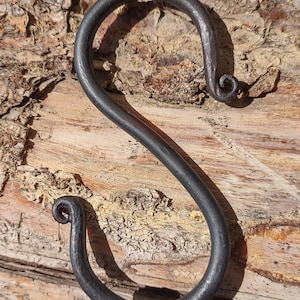 Pode incluir: Gancho em S de metal cinza escuro forjado à mão com extremidades enroladas. O gancho é exibido contra um fundo de madeira castanha clara texturizada. Este gancho decorativo é adequado para pendurar itens.
