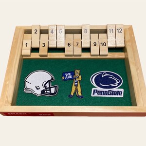 May include: A wooden game board with a green felt surface and a set of numbered tiles. The board also features three embroidered patches: a football helmet, a cartoon bear holding a sign that says "We Are", and a Penn State Nittany Lion logo. The board is labeled "Smash Box".