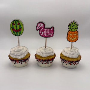May include: Three cupcakes with white frosting in patterned liners, each topped with a decorative pick. The picks feature a green watermelon jack-o'-lantern, a pink flamingo skeleton, and an orange pineapple jack-o'-lantern on thin sticks.