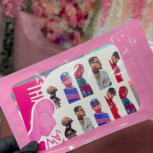 May include: A pink package containing a sheet of stickers with images of a person wearing hats, sunglasses, and various outfits. The stickers have the word "TEMENTS" on them. The package is held by a gloved hand.
