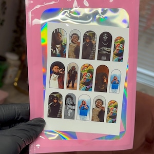 May include: A pink holographic pouch containing a sheet of fifteen rectangular nail decals. Each decal features a distinct portrait or artistic graphic of a person, arranged in three rows of five, with rounded top edges on each individual sticker.