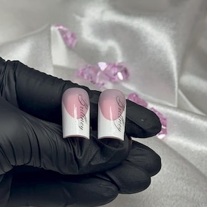 May include: Two artificial nails with a white base and a pink gradient, each bearing the name "Britney" in elegant script. The nails are displayed against a backdrop of white fabric and pink crystals.